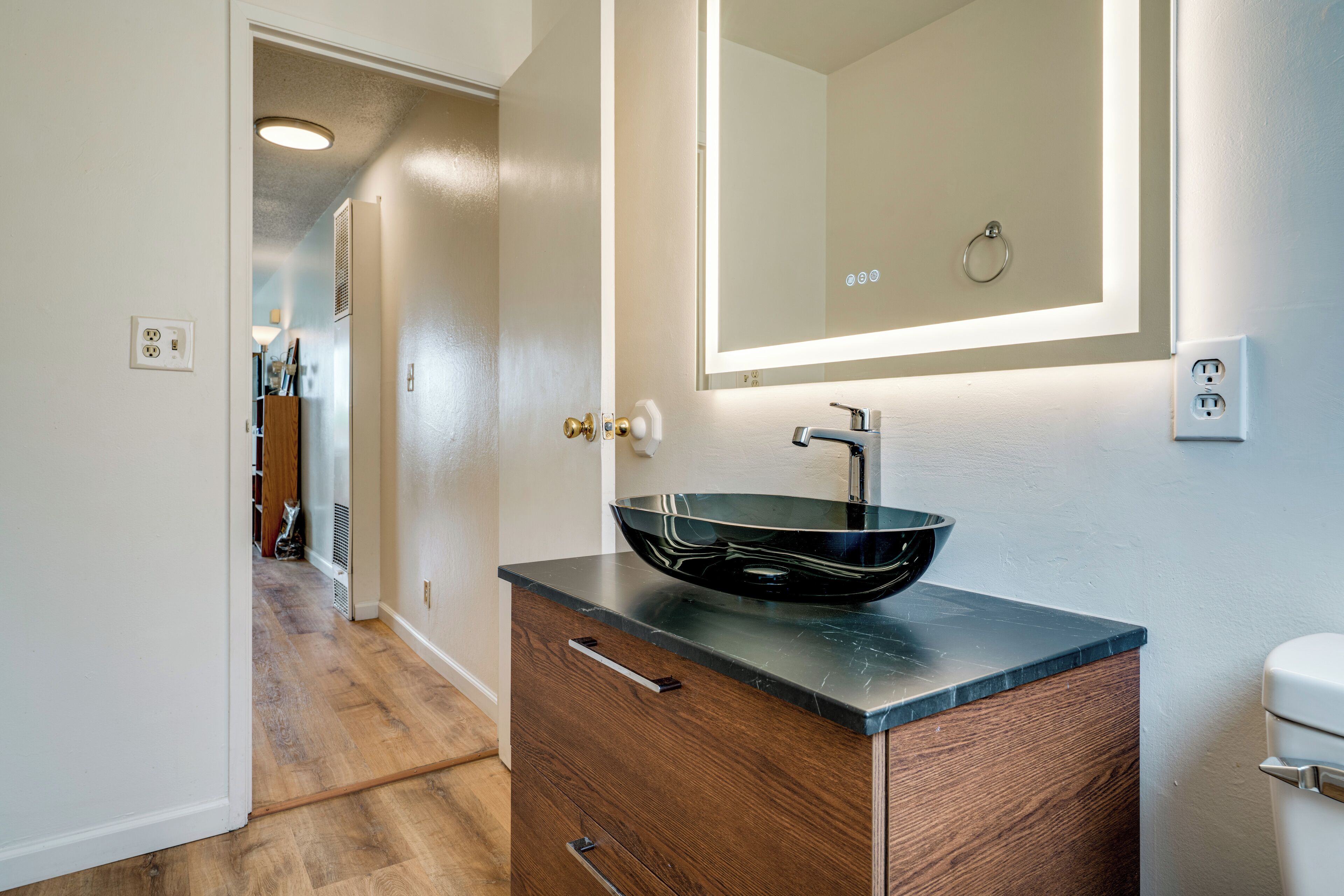 Un baño con un lavamanos tipo cuenco negro, un mueble de madera y un espejo con iluminación integrada.