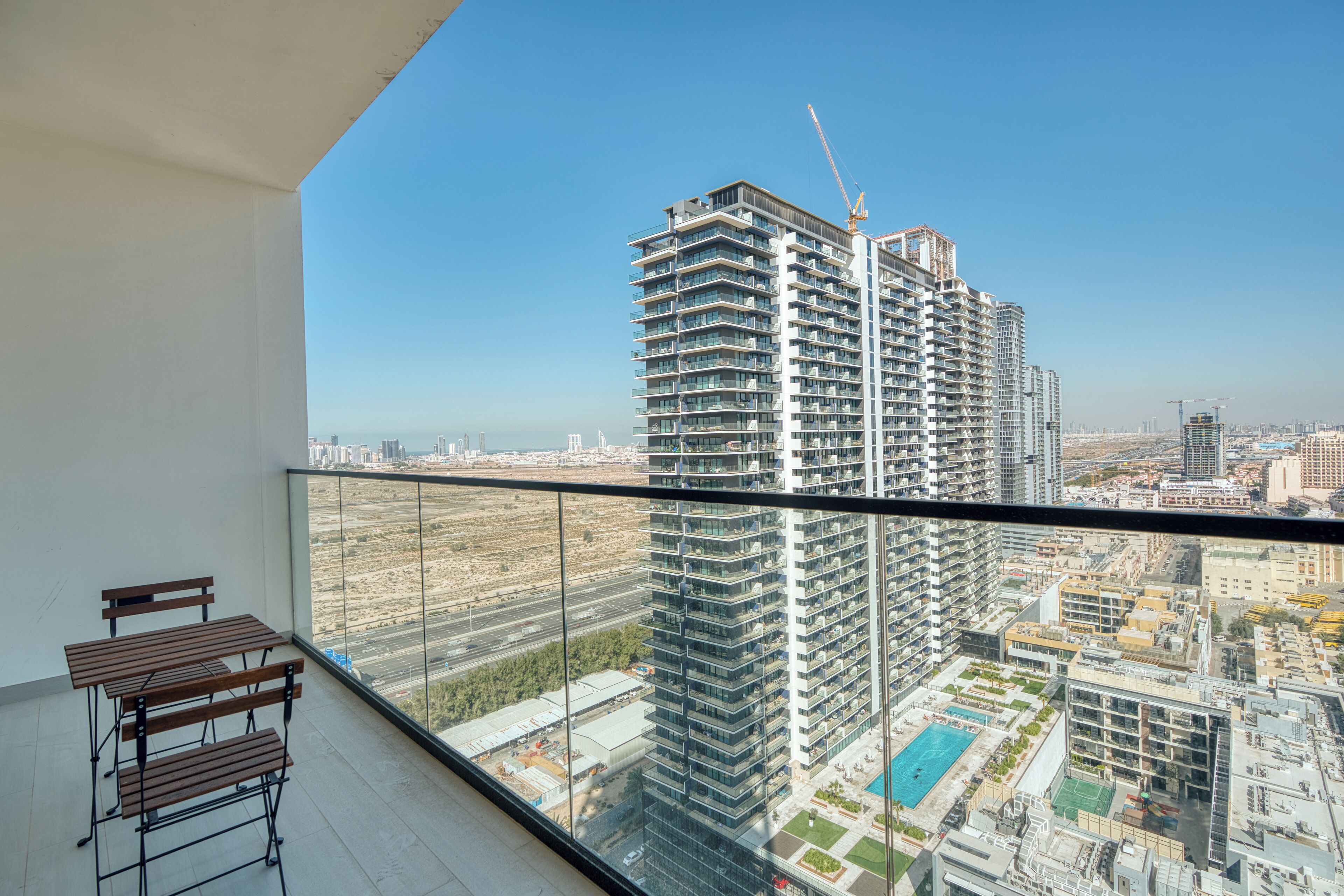 A high-rise building with a construction crane, a balcony with a wooden table and bench, and a view of a cityscape.