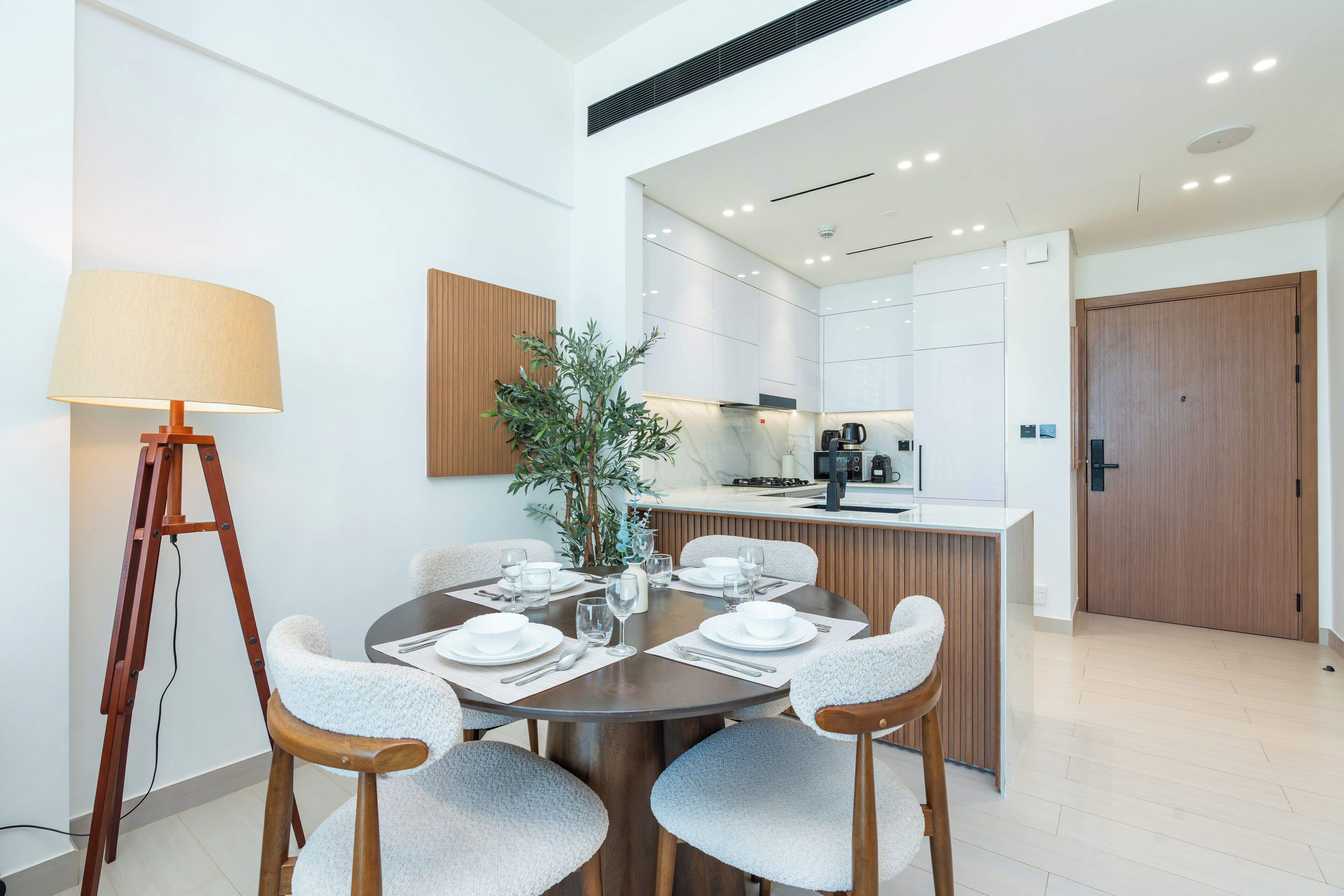 A modern dining area with a round table set for four, a wooden side table with a lamp, and a kitchen in the background.