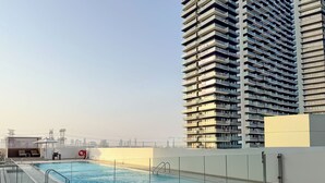 Een zwembad op het dak met een glazen balustrade, een loungegedeelte met stoelen, en een hoog gebouw op de achtergrond.