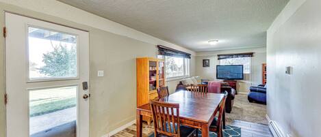 A dining area with a wooden table and chairs, a bookshelf, and a television in a living room with a view of the outdoors.