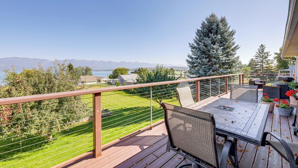 A deck with a table and chairs, overlooking a green lawn and a mountain range. - Hot Tub, Views & Private Dock: Polson Haven! (Polson)