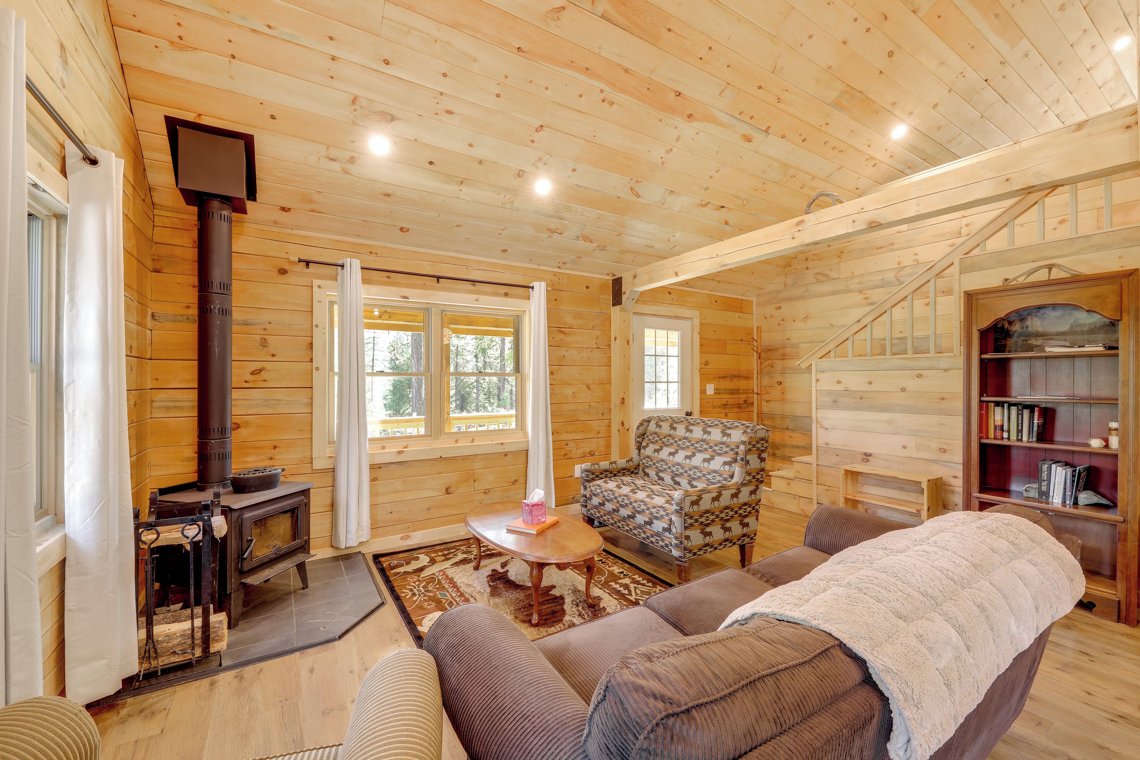 Acogedor interior de cabaña de madera con chimenea, un sofá, un sillón estampado y una estantería.
