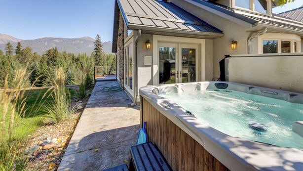 Un bain à remous dans un jardin, avec vue sur les montagnes, une terrasse en bois et une maison dotée de grandes fenêtres et d’un toit à pente.