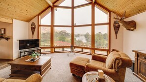 A spacious living room with a large wooden ceiling, a mounted deer head, a television, and a view of a lake through large windows. - Stunning A-frame Home w/ Views, 10 Mi to Okanogan! (Brewster)
