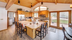 A spacious kitchen with wooden cabinets, a central island with chairs, and a view to the outside. - Stunning A-frame Home w/ Views, 10 Mi to Okanogan! (Brewster)