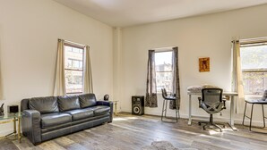 Ein Wohnzimmer mit einem Ledersofa, einem Glastisch, einem schwarzen Bürostuhl und einem kleinen weißen Tisch mit einer Lampe.