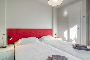 Quarto com duas camas, uma cabeceira vermelha, abajures e uma toalha dobrada na cama à direita.