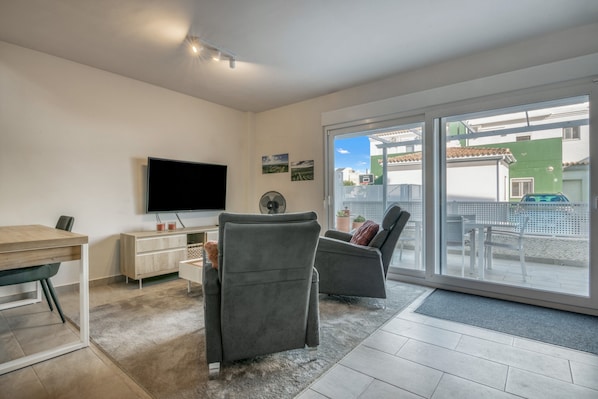 Uma sala de estar moderna com televisão de tela plana, duas poltronas cinzas, uma mesa de jantar de madeira e uma grande porta de vidro deslizante que dá para uma área externa.