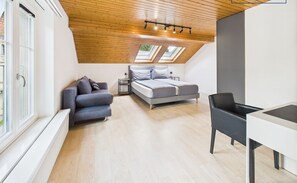 A modern bedroom with a sloped ceiling, skylights, a bed, a sofa, a desk, and a chair.