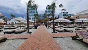 Un hotel frente a la playa con un camino central que lleva a un edificio, rodeado de tumbonas y palmeras.