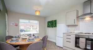 A compact kitchen with a dining area, a white refrigerator, and a window with blinds.