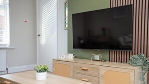 A room with a large flat-screen TV mounted on the wall, a wooden cabinet with drawers, a small table with a potted plant, and a calendar on the cabinet.