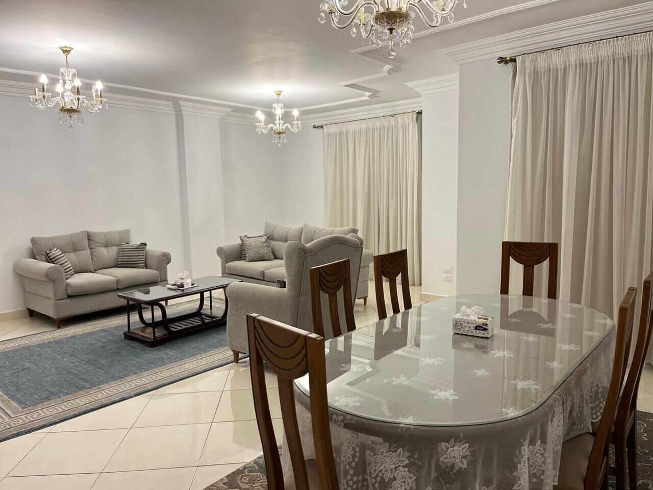 Una sala de estar moderna con una mesa de comedor de vidrio, dos sofás, sillas de madera, una mesa baja y una araña.