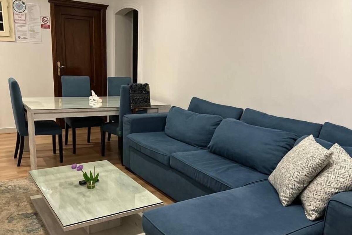 Ein Wohnzimmer mit einem blauen Ecksofa, einem Glastisch und einem Essbereich mit Tisch und Stühlen.