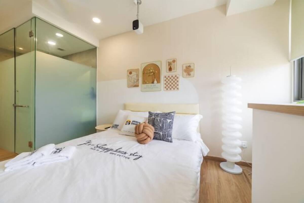 A hotel room with a bed, a glass shower enclosure, a wall-mounted light fixture, and artwork.