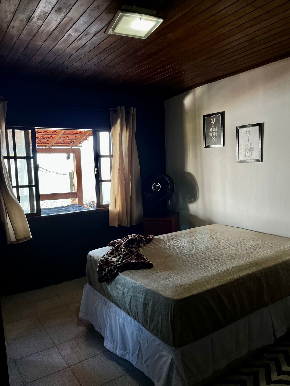 Une chambre à coucher avec un lit, une fenêtre avec des rideaux, un ventilateur fixé au mur et deux tableaux encadrés.