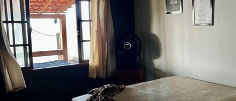 Une chambre à coucher avec un lit, une fenêtre avec des rideaux, un ventilateur fixé au mur et deux tableaux encadrés.