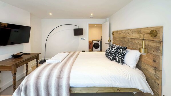 A modern bedroom with a large bed, a flat-screen TV, a desk, and a washing machine in the background.