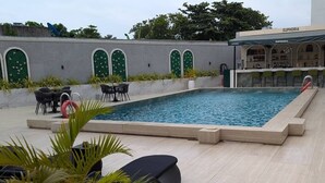 Ein Swimmingpool mit Liegestühlen und Tischen, umgeben von einer niedrigen Mauer mit Rundbogenfenstern.