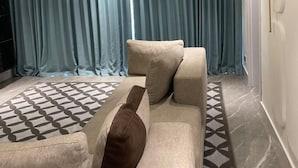 Ein stilvolles Wohnzimmer mit einem beigefarbenen Sofa, einem gemusterten Teppich und einem großen Fenster mit blauen Vorhängen.