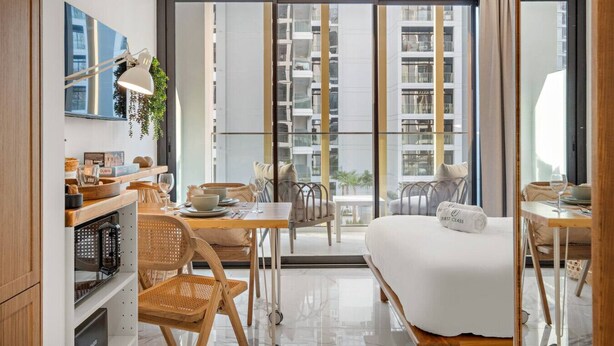 A modern hotel room with a large sliding glass door to a balcony, a bed, a desk with a lamp, and a view of the cityscape.