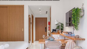 A hotel room with a bed, a dining table with chairs, a TV, a plant, and a shelf with items.