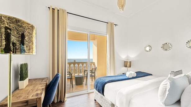 Une chambre d’hôtel comprenant un lit, un bureau, une chaise, une plante en pot, un balcon avec vue et des miroirs décoratifs.