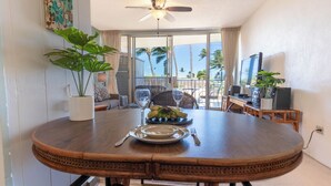 Een eethoek met een ronde houten tafel, stoelen, een plafondventilator en uitzicht op palmbomen door grote ramen.