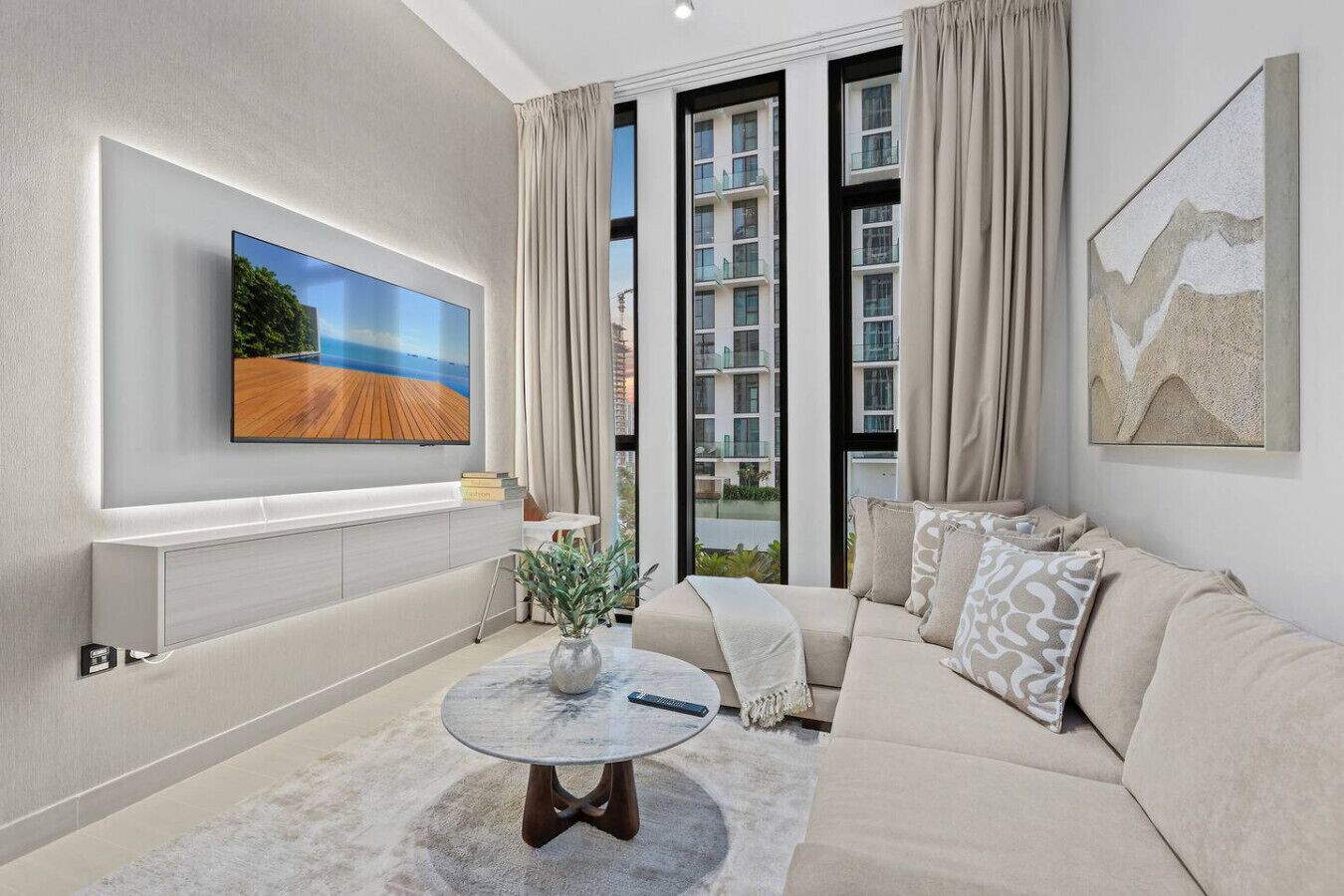 A modern living room with a large TV, a sofa, a coffee table, and a view of a building through large windows.