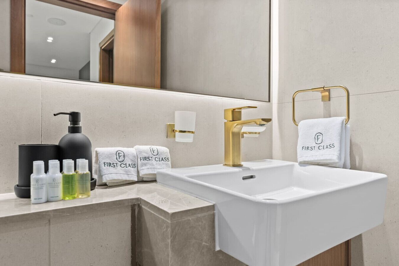 A modern bathroom with a white sink, gold faucet, and a towel rack with 'First Class' towels. There are also black soap dispensers and bottles on the counter.