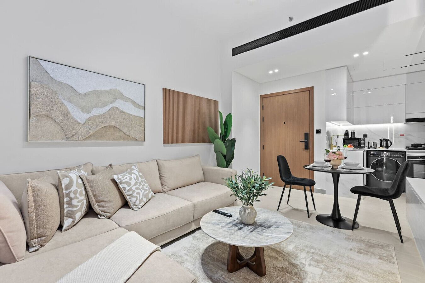 A modern living room with a sofa, a coffee table, and a dining area with chairs. There is a kitchen in the background with white cabinets and appliances.