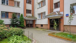 A multi-story residential building with a brick facade, multiple windows, and a small garden area with plants and a wooden planter. - Spacious Apartment | Air Conditioning, Balcony, close to the Stadium (Warszawa)