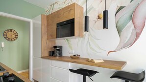 Eine moderne Küche mit einer Holzausziehtheke, einem eingebauten Backofen und einer Kaffeemaschine. Zwei schwarze Barhocker und eine Wanduhr sind ebenfalls vorhanden.