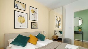 Ein ordentlich bezogenes Bett mit einem grün-gelben Kissen, einer weißen Decke und einem gefalteten Handtuch. An der Wand hängen gerahmte Kunstwerke und über dem Bett befindet sich ein Spiegel.