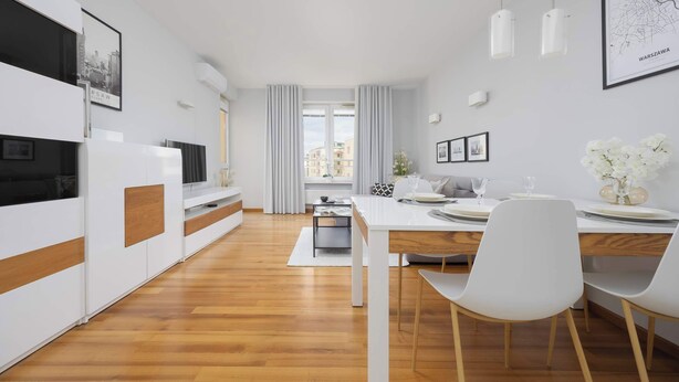 Una sala de estar moderna y bien iluminada con una mesa de comedor para dos personas, un sofá y un televisor sobre un mueble blanco.