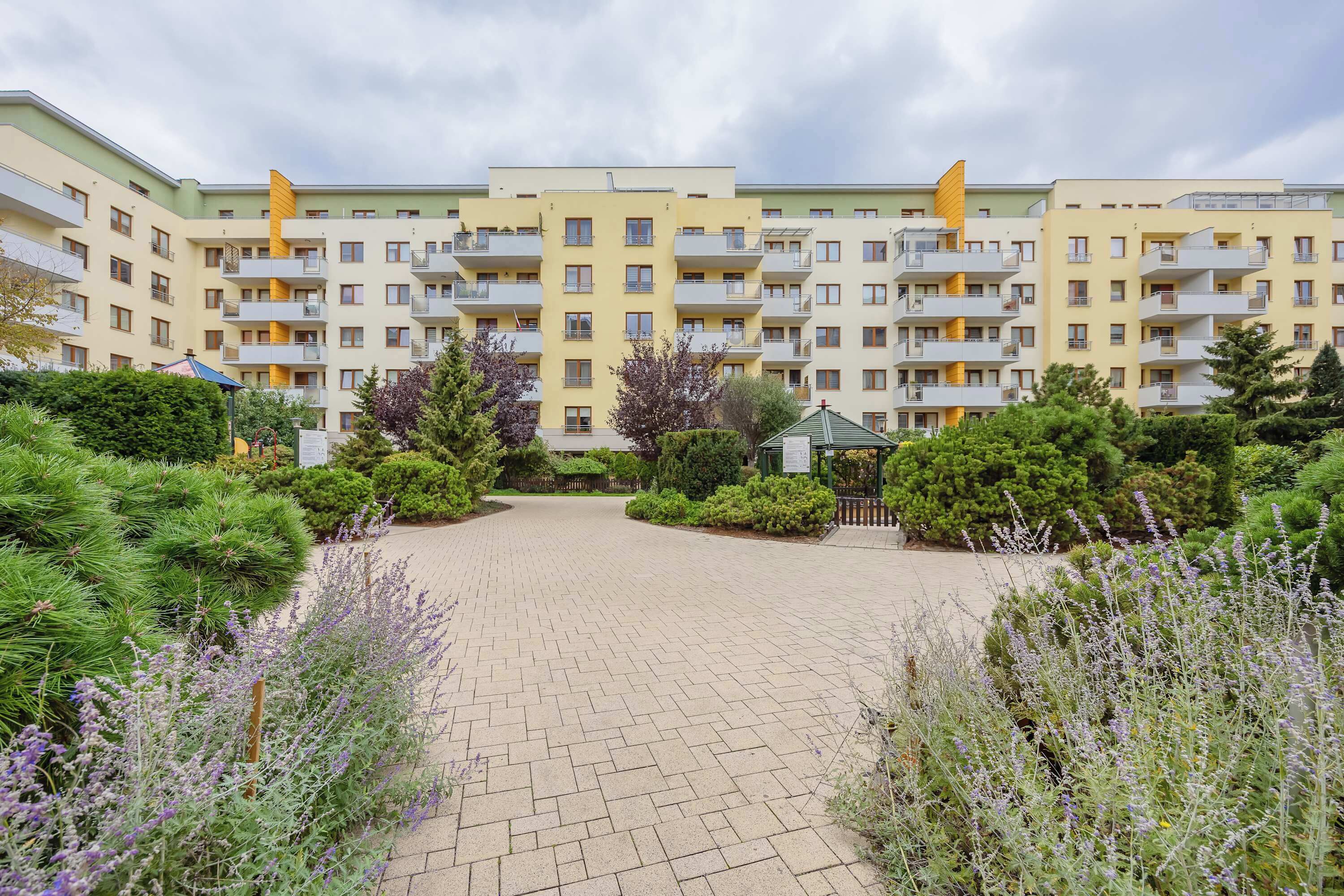 A multi-story residential building with balconies, surrounded by well-maintained gardens and a paved courtyard.