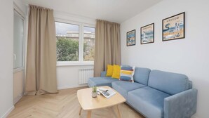 Un salón con un sofá azul, una mesa de centro de madera y una ventana con cortinas. Hay dos cuadros en la pared.
