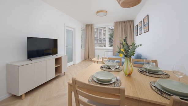 Un comedor moderno con una mesa de madera dispuesta para dos, un televisor sobre un mueble blanco, y un balcón con vistas a edificios.