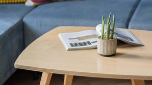 Una sala de estar moderna con una mesita de centro de madera clara, una pequeña planta en maceta y una revista. Un sofá azul con almohadones coloridos se encuentra al fondo.