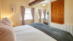 A bedroom with a bed, floral curtains, a wooden ceiling beam, and a radiator. - Host & Stay | Dairy Cottages (Masham)