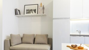 A modern living room with a beige sofa, a small kitchen area with a white countertop, and a shelf with books and a framed picture. - 7Studios - 7 Blue (Milano)