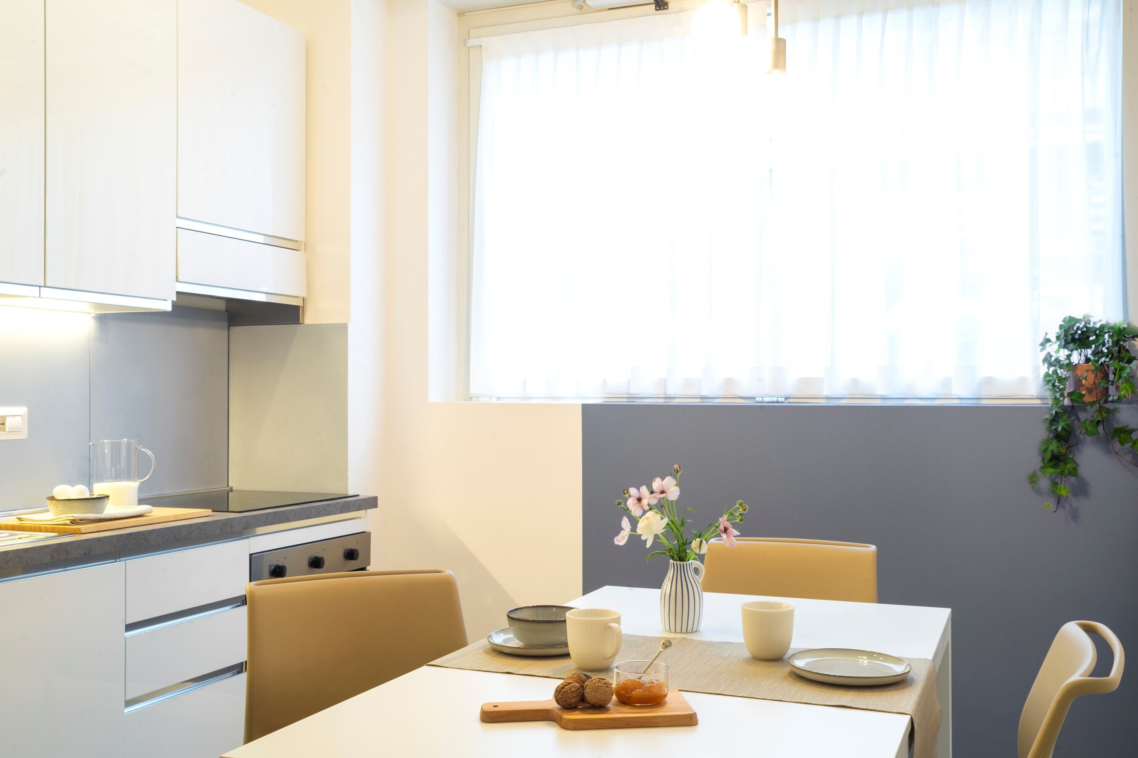 A modern kitchen with a dining table set for two, featuring a vase of flowers, a cutting board with nuts, and a bowl of fruit.