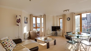 A modern living room with a sofa, coffee table, TV, and dining area with chairs. - Harbourside Apartment (Weymouth)