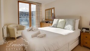 A bedroom with a large bed, a chair, a nightstand, and a window with curtains. - Harbourside Apartment (Weymouth)