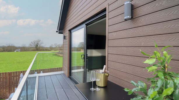 A balcony with a wooden floor, a table with glasses and a bottle, and a view of a green field.