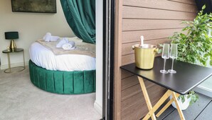 A bedroom with a bed, green curtains, a bedside lamp, and a small table with a champagne bottle and glasses.