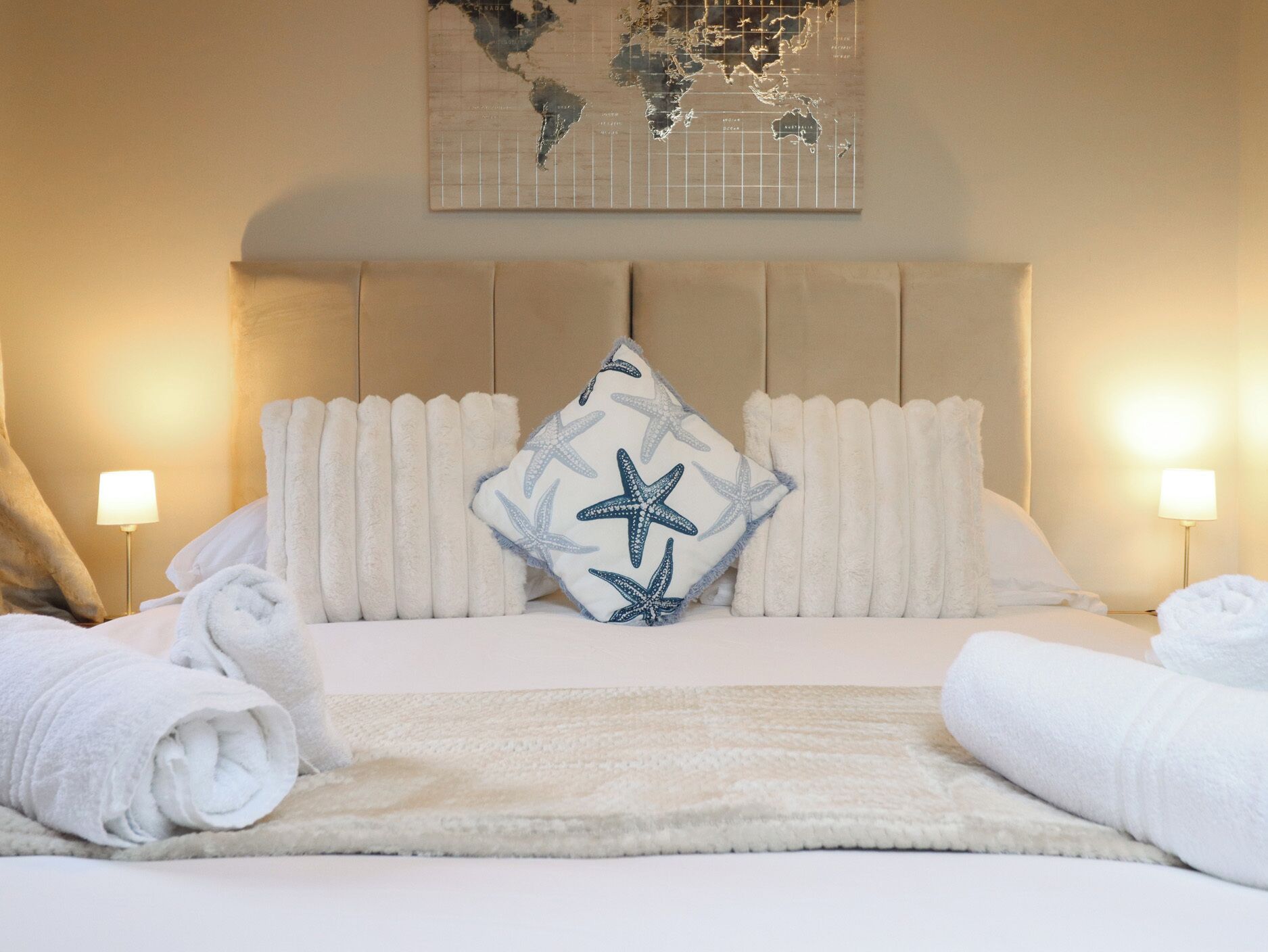 Un lit bien fait, avec du linge de lit blanc, un coussin en forme d’étoile de mer et deux lampes de chaque côté.