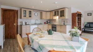 A compact kitchen with wooden cabinets, a dining table set with a green tablecloth, and a small potted plant. - Bluebell Cottage (Tenbury Wells)