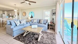 A living room with a sectional sofa, a coffee table, and a patterned rug. There is a ceiling fan, a kitchen in the background, and a view through large windows. - Summer & July 4th Savings at Beachside Colony Tybee's ONLY Oceanfront Resort 2/2 (Tybee Island)